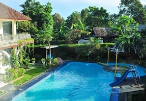 A swimming pool with a slide, surrounded by lush greenery and a thatched-roof structure. - The Garden Family Guest House (Ciawi)
