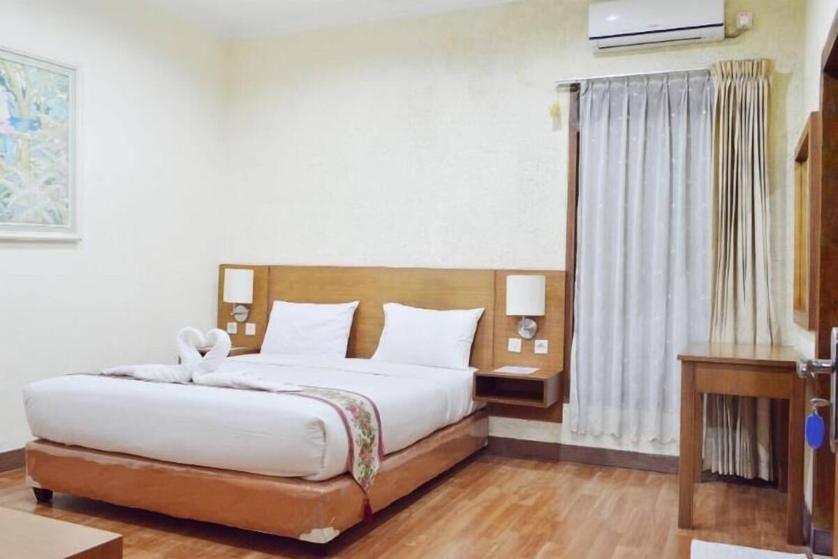 A hotel room with a wooden bed, white linens, a nightstand, a lamp, an air conditioner, and a window with curtains.