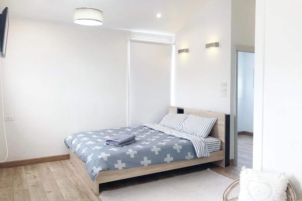 A modern bedroom with a wooden bed, a grey and white patterned bedspread, and a bedside table with a lamp.