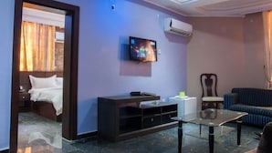 A hotel room with a bed, a TV mounted on the wall, a sofa, a glass coffee table, and an air conditioning unit. - Kapino Suites (Abuja)