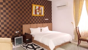 A hotel room with a checkered wall, a bed with white bedding, a small bedside table, a chair, and a picture frame. - Kapino Suites (Abuja)