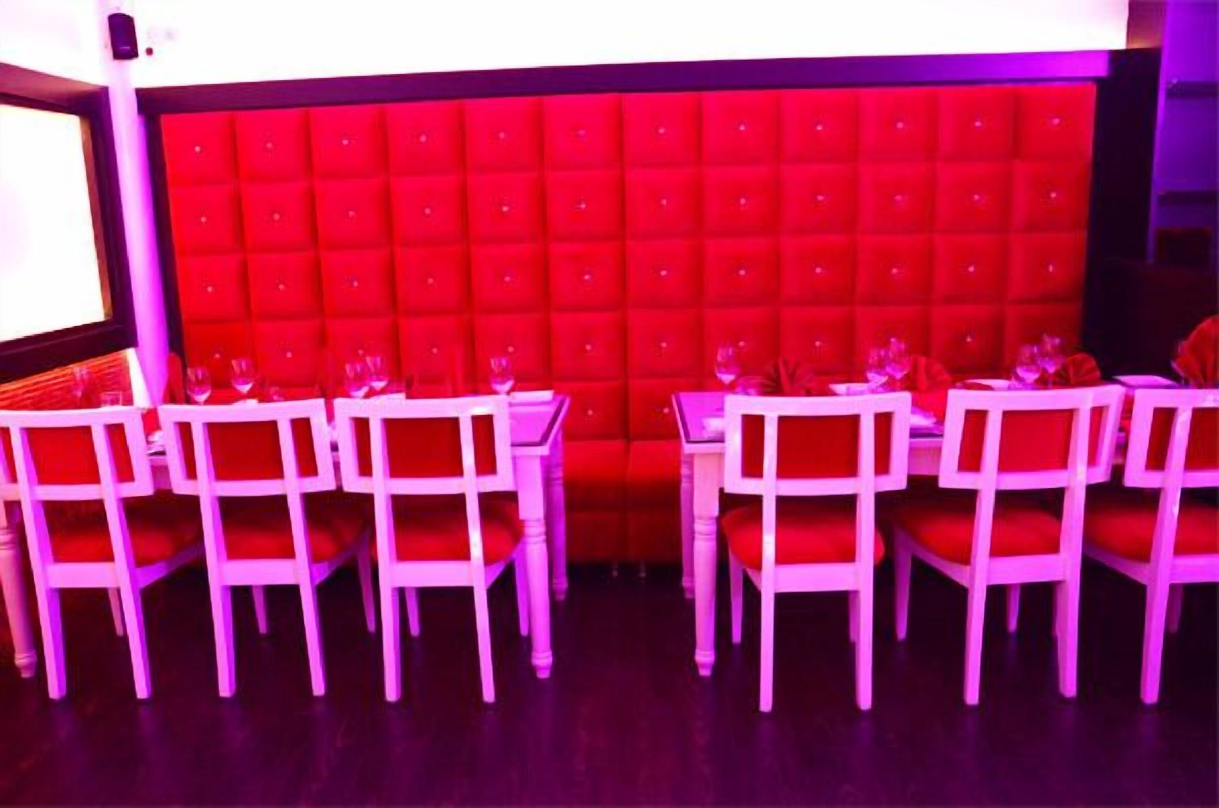 A dining area with white tables and chairs, a red upholstered wall, and a screen on the left.