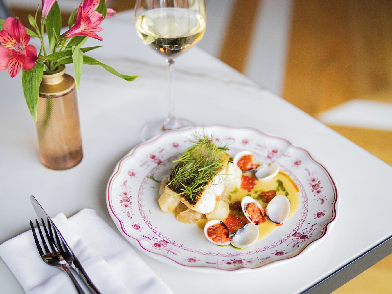 Taula de dinar amb un plat de menjar, un got de vi blanc i un ram de flors.