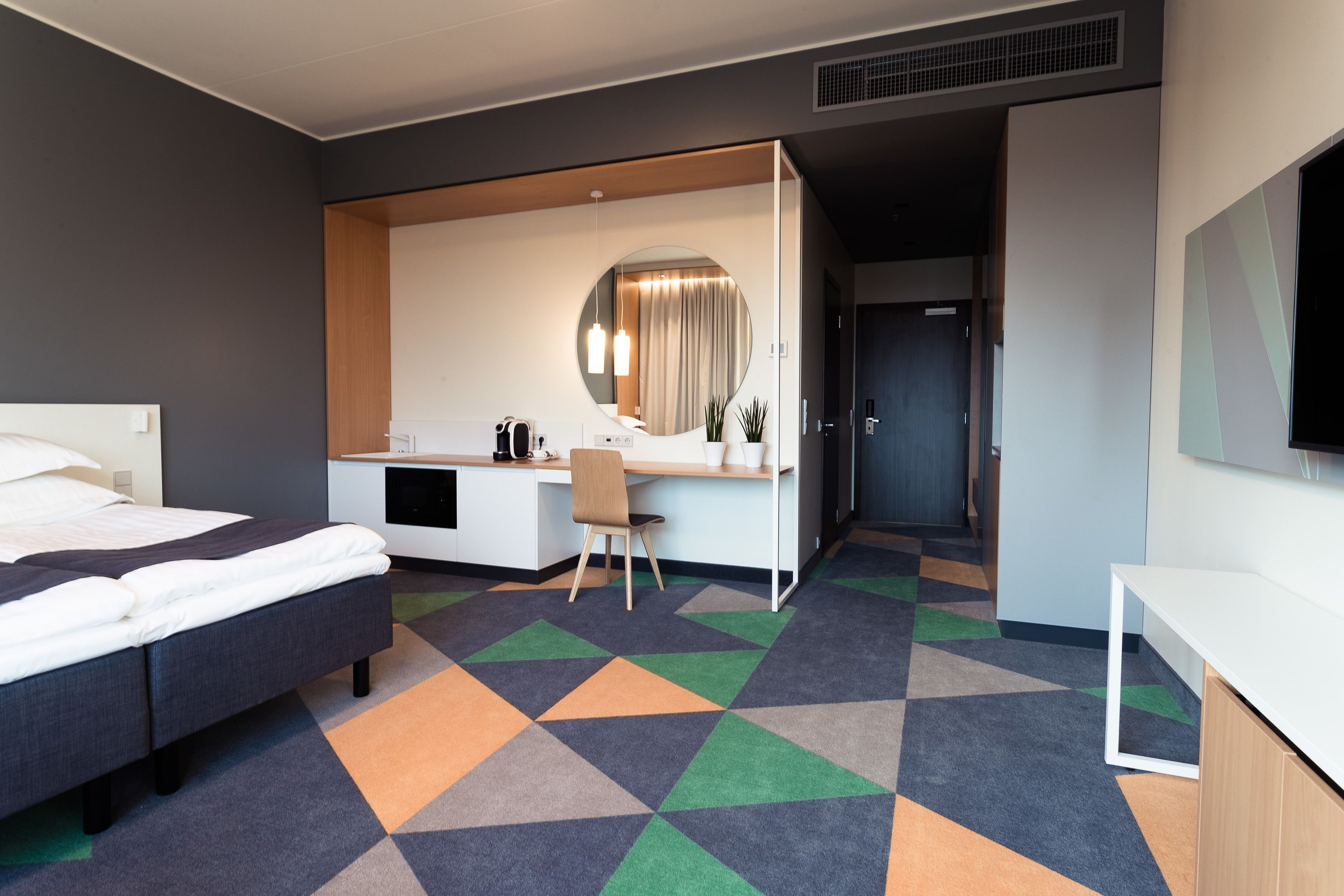 A modern hotel room with a bed, desk, chair, and a large mirror.