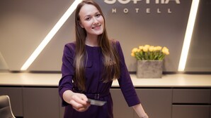 Una mujer con un vestido morado está de pie frente a la mesa de recepción, donde se lee el letrero "Sophia Hotel".