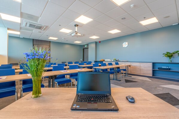 Un aula con una laptop sobre un escritorio, sillas azules y un florero con flores.