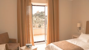 A hotel room with a bed, a chair, a small table with books, and a view of a hillside through an open door. - Hotel Solar do Bom Jesus (Santa Cruz)