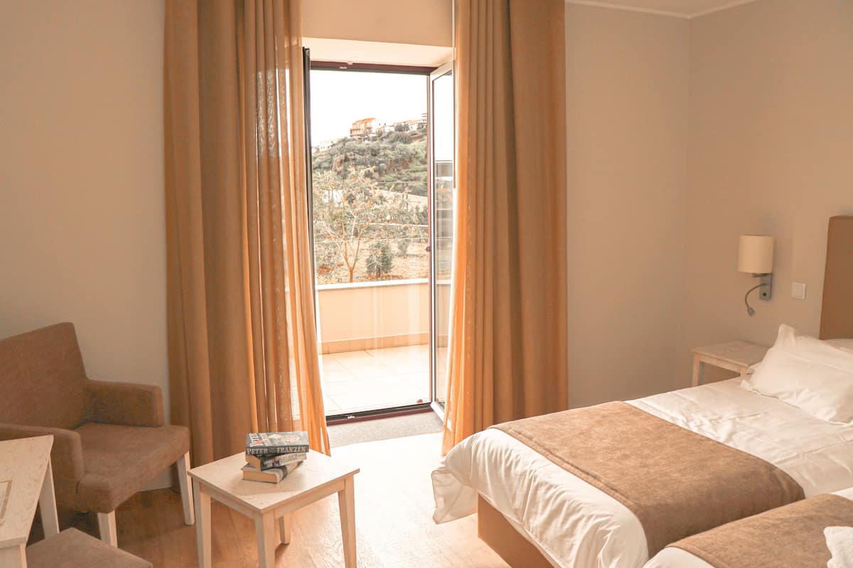 A hotel room with a bed, a chair, a small table with books, and a view of a hillside through an open door.