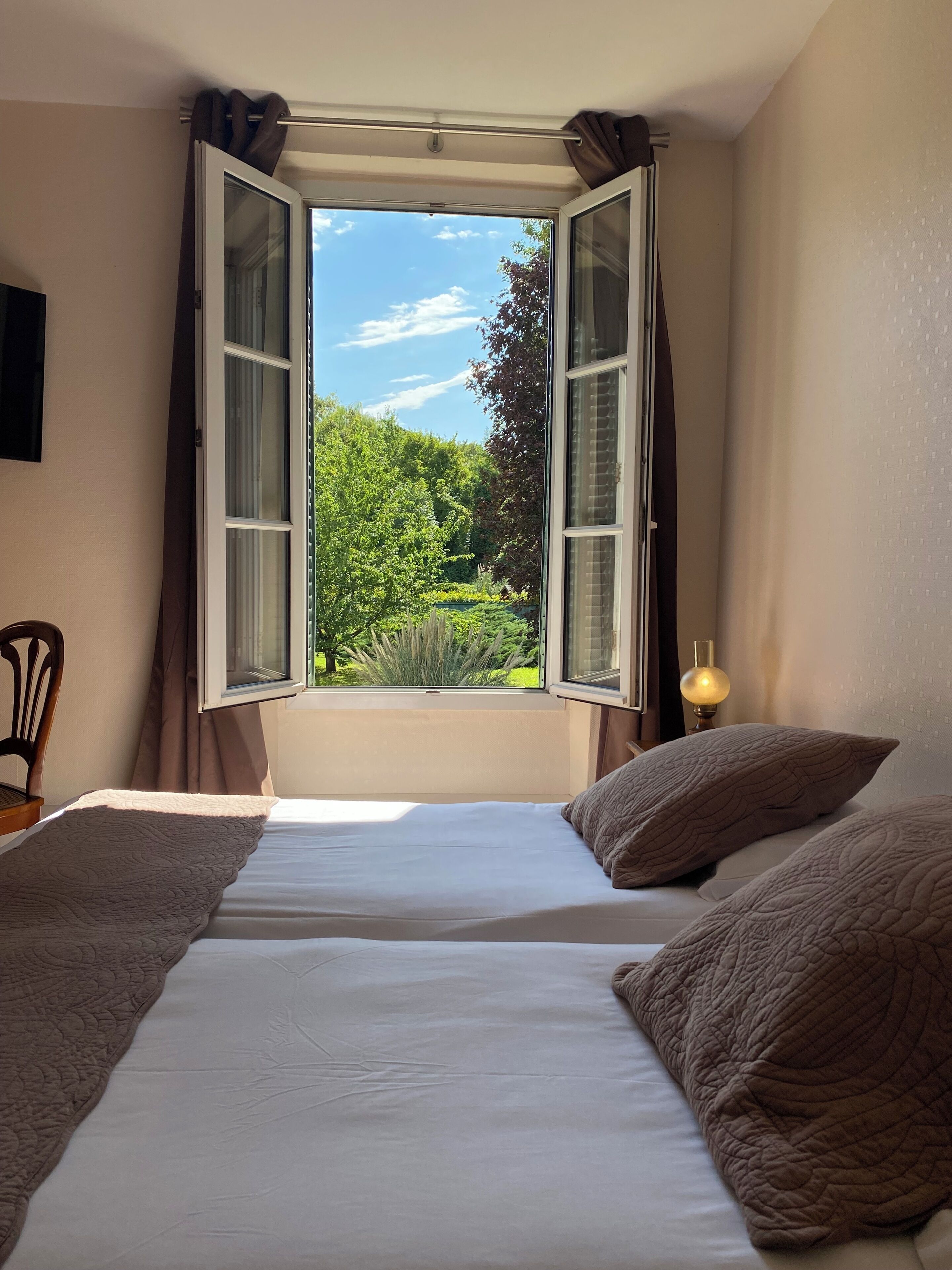 Een slaapkamer met een open raam, een bed met kussens, een stoel en een bedlamp.
