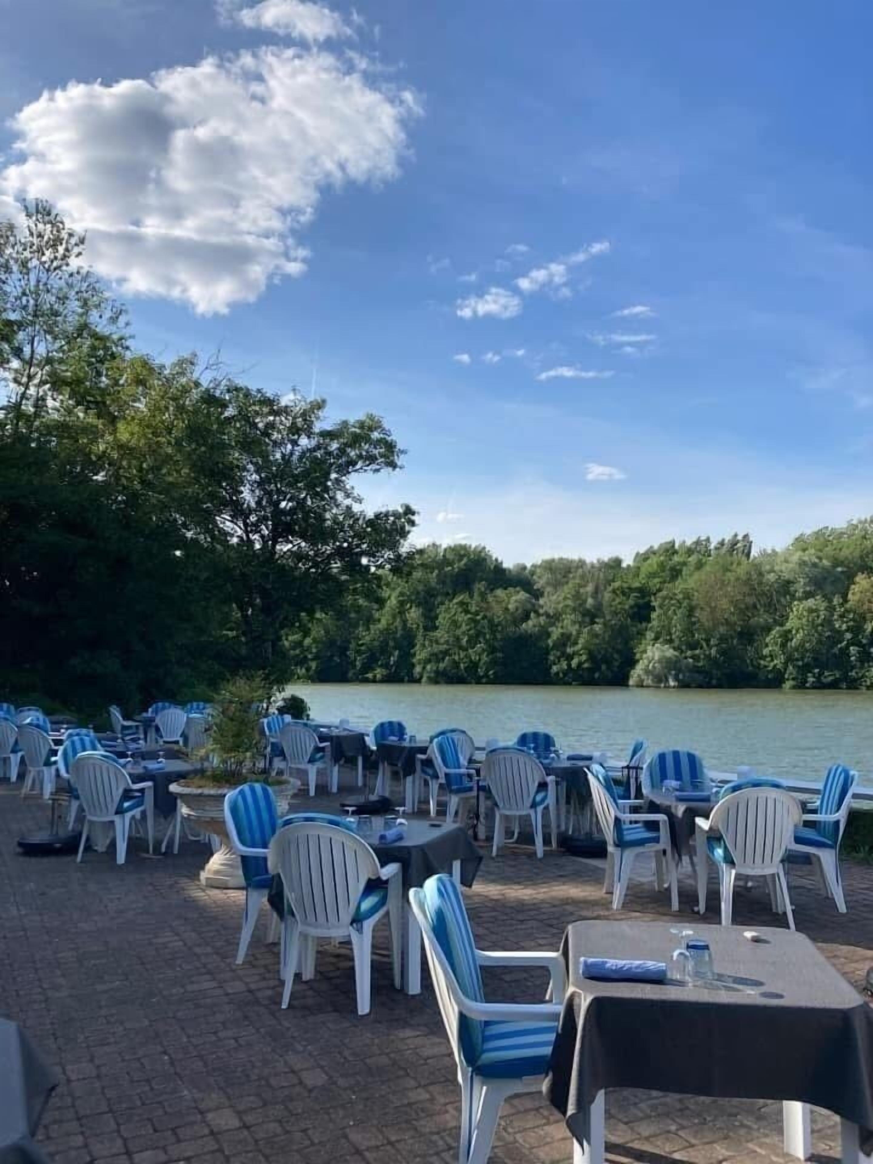 Outdoor seating with tables and chairs, a body of water, and trees in the background.
