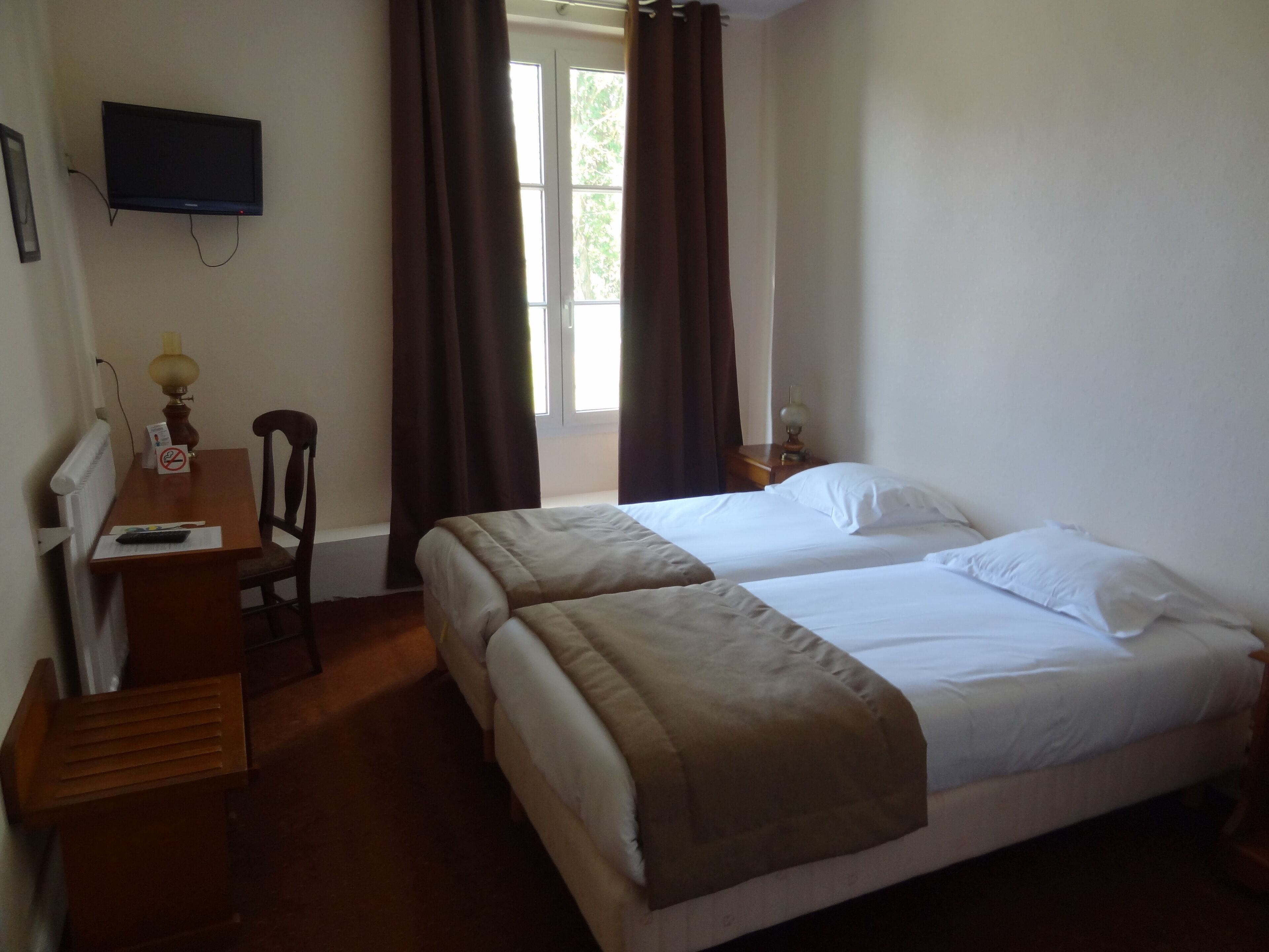 A hotel room with a bed, a desk, a chair, a TV mounted on the wall, and a window with curtains.