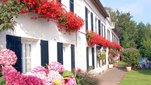 Un edificio blanco con contraventanas negras, un sendero de ladrillo y coloridas flores rosas y rojas en primer plano.
