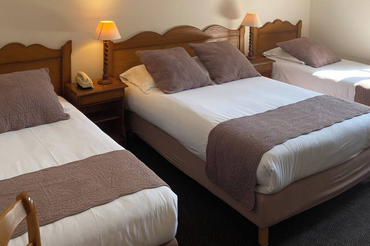 A hotel room with three beds, each with a wooden headboard, white linens, and a brown throw blanket. There are bedside lamps and a telephone on the nightstands.