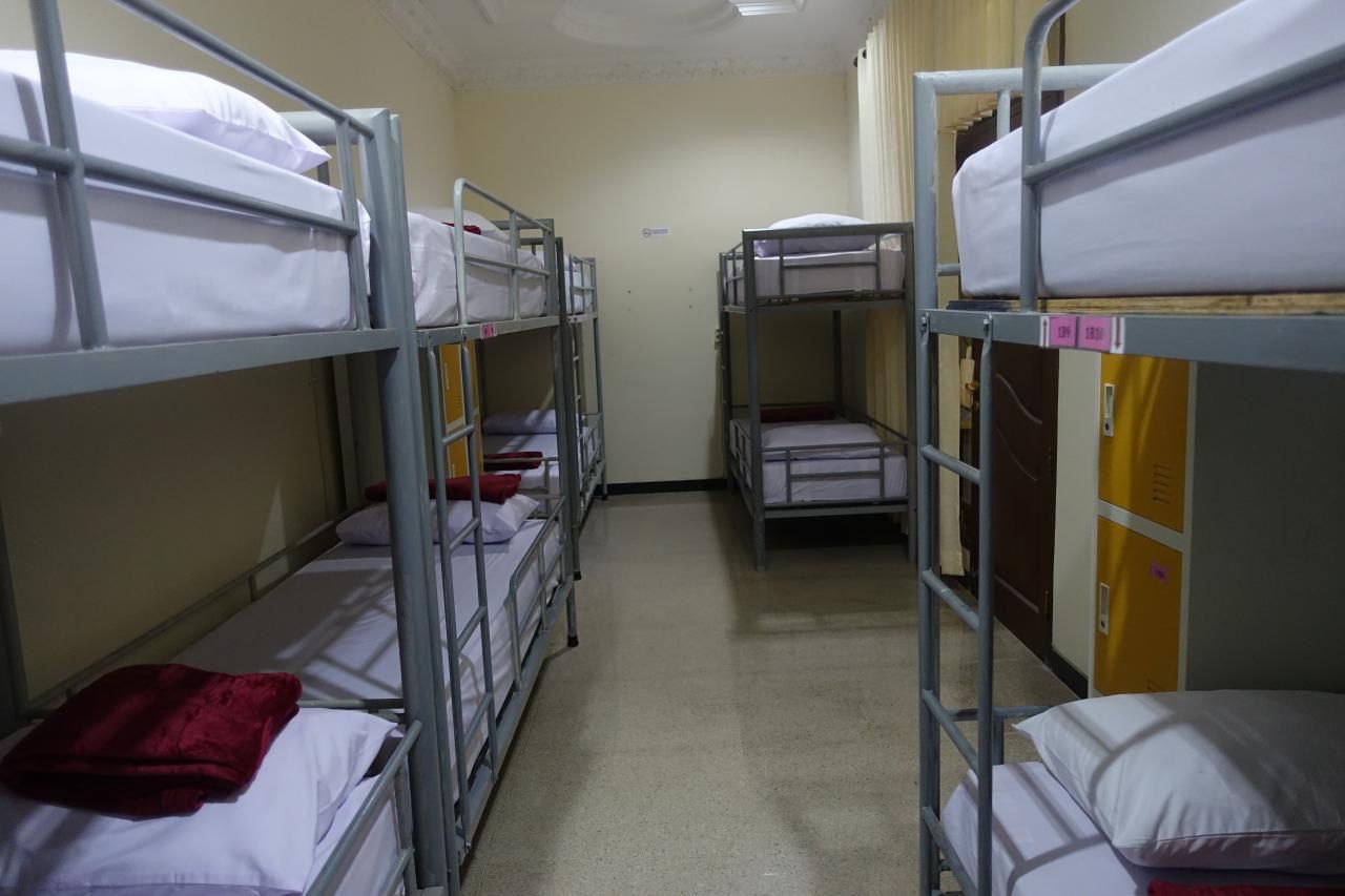 Un pasillo con literas dispuestas en dos filas, cada una con colchón y almohadas.