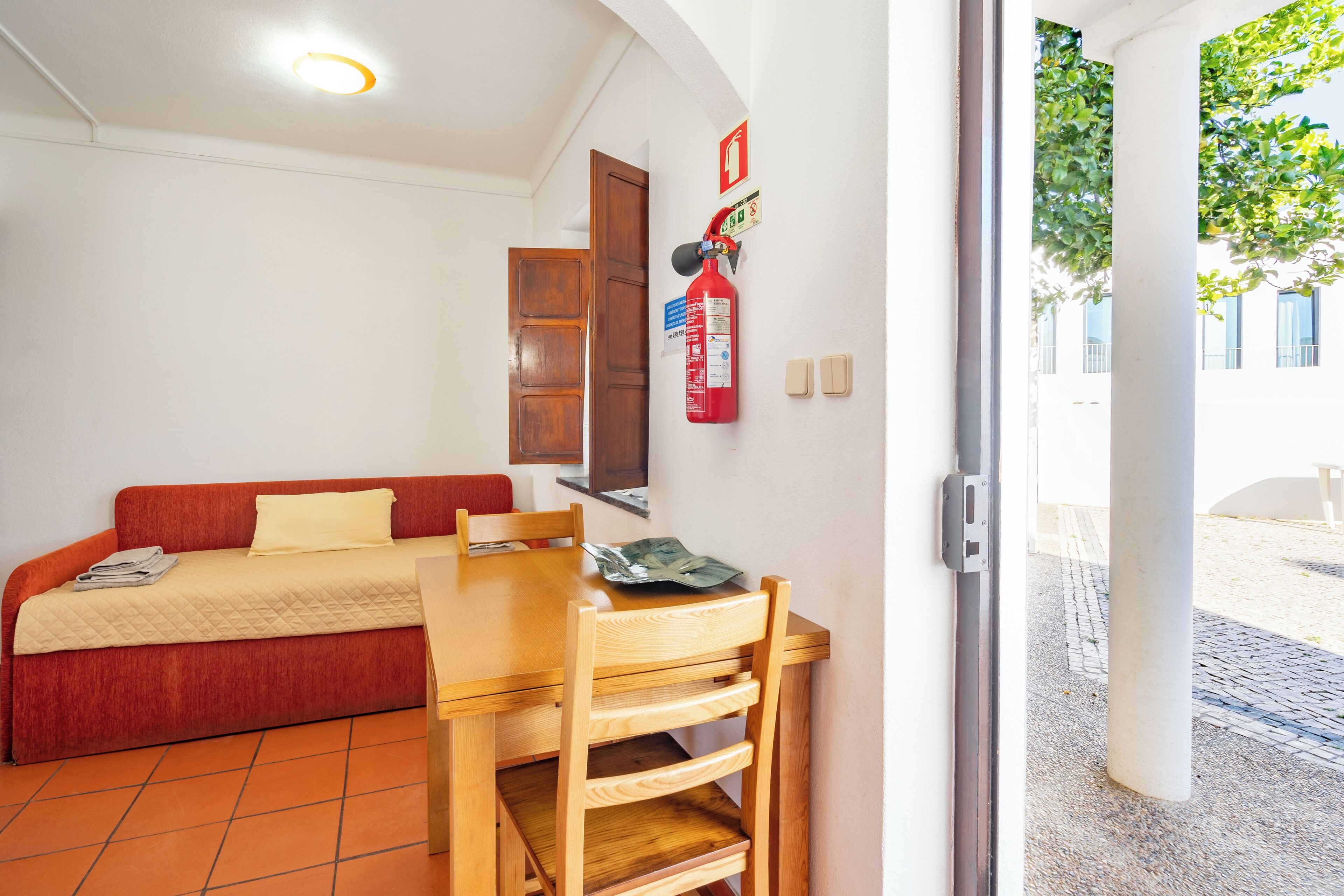 Una habitación con una cama, una mesa de madera y una silla. Hay un extintor en la pared y una puerta que da al exterior.