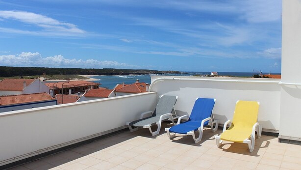 Un balcón con tres sillones de descanso, piso de baldosas y vista al mar y edificios.