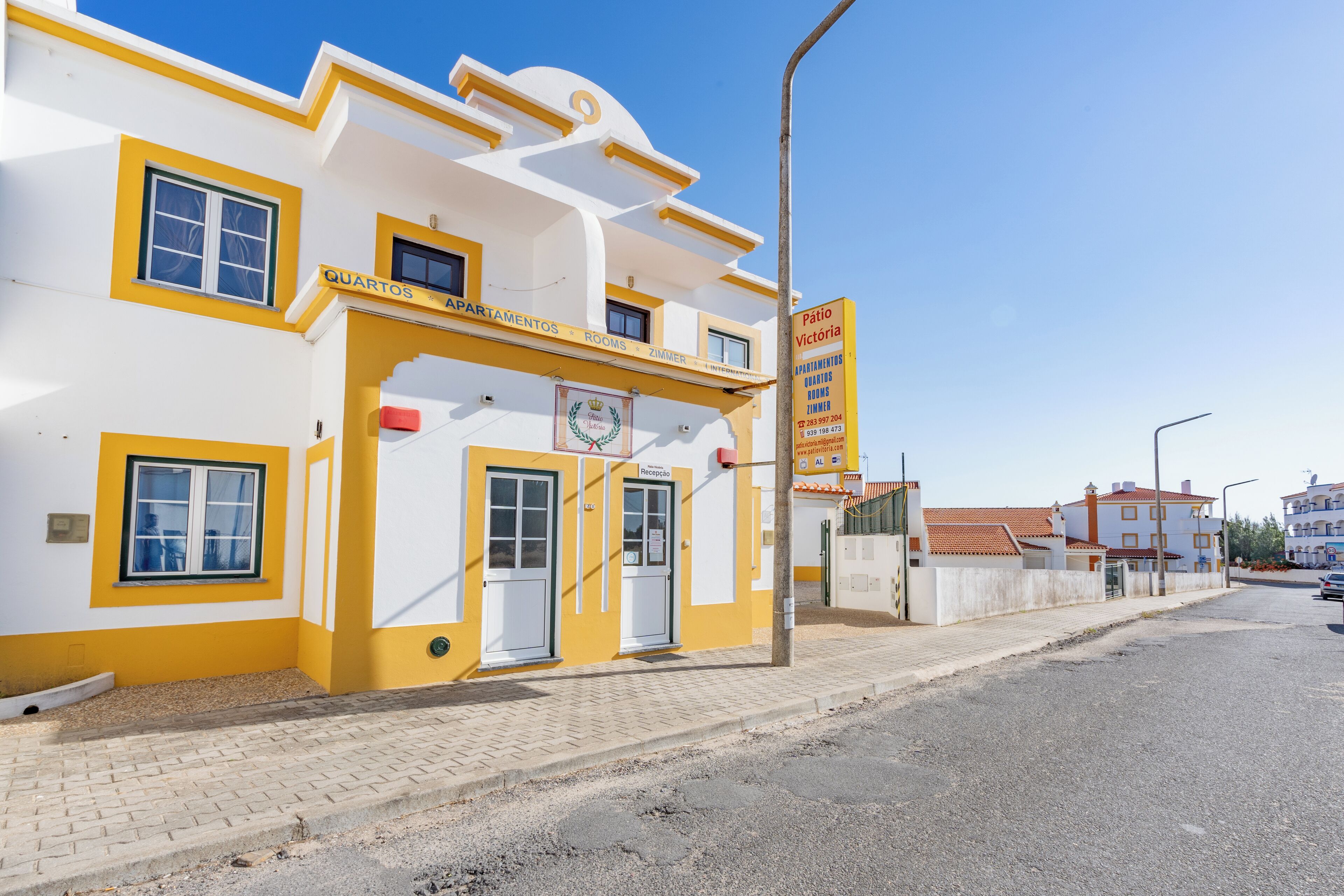 A building with a yellow and white facade, a signboard for 'Pátio Vitória Apartments,' and a clear blue sky.