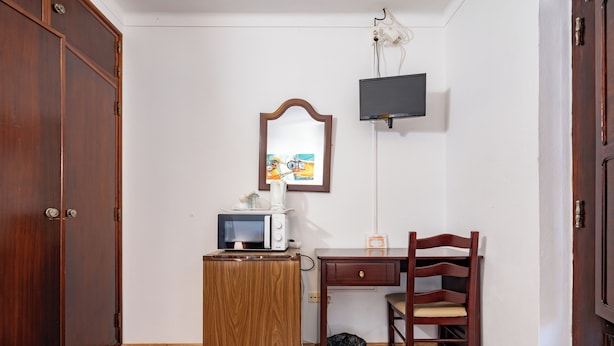 A compact room with a wooden wardrobe, a small desk with a chair, a microwave, and a TV mounted on the wall.