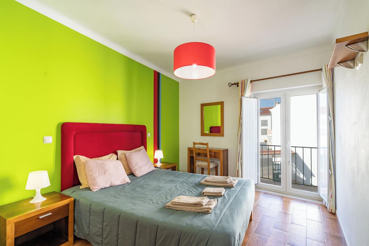Un dormitorio con cama, mesita de noche, lámpara y vistas al balcón exterior.