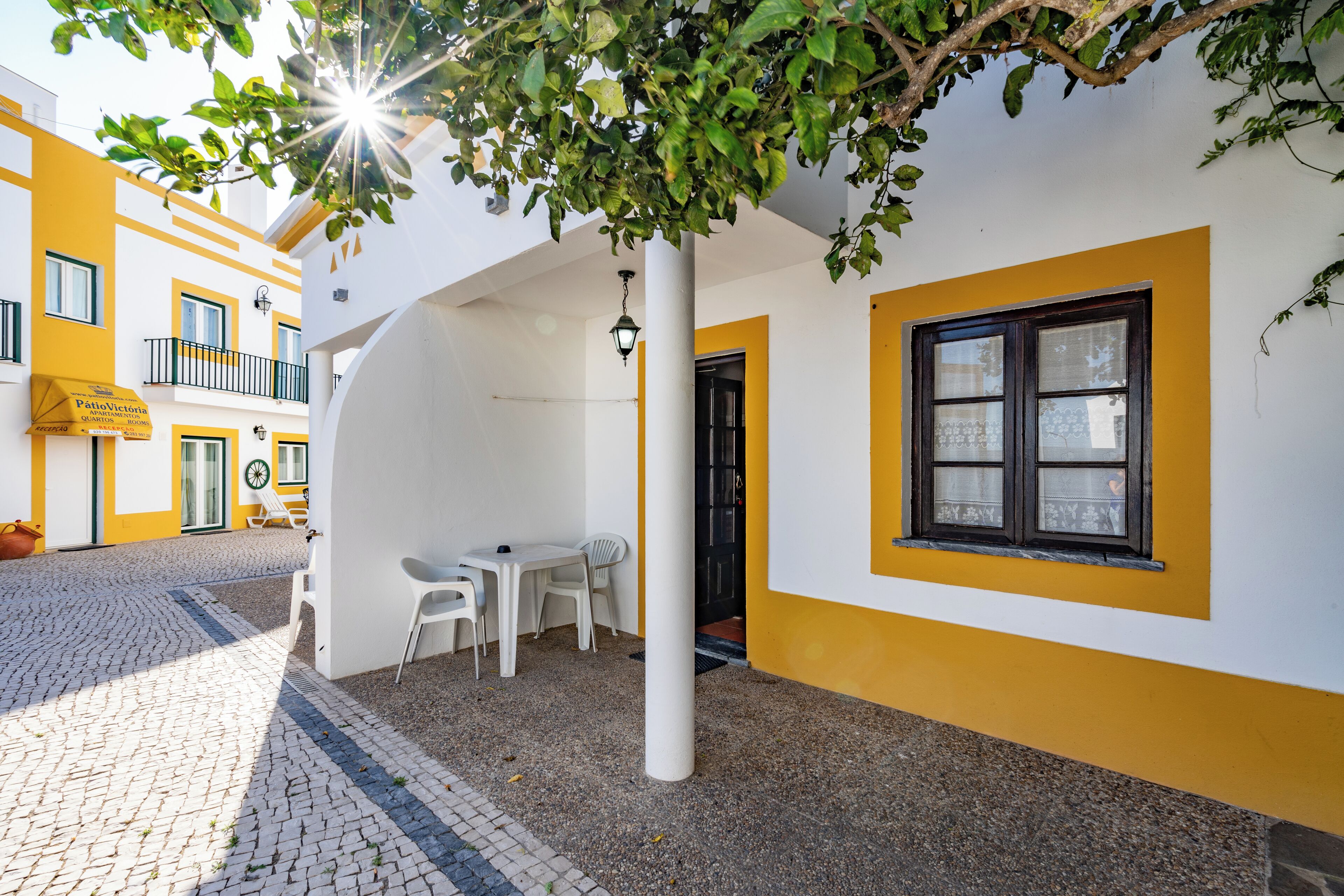 A courtyard with a white table and chairs, a yellow and white building, and a tree providing shade.