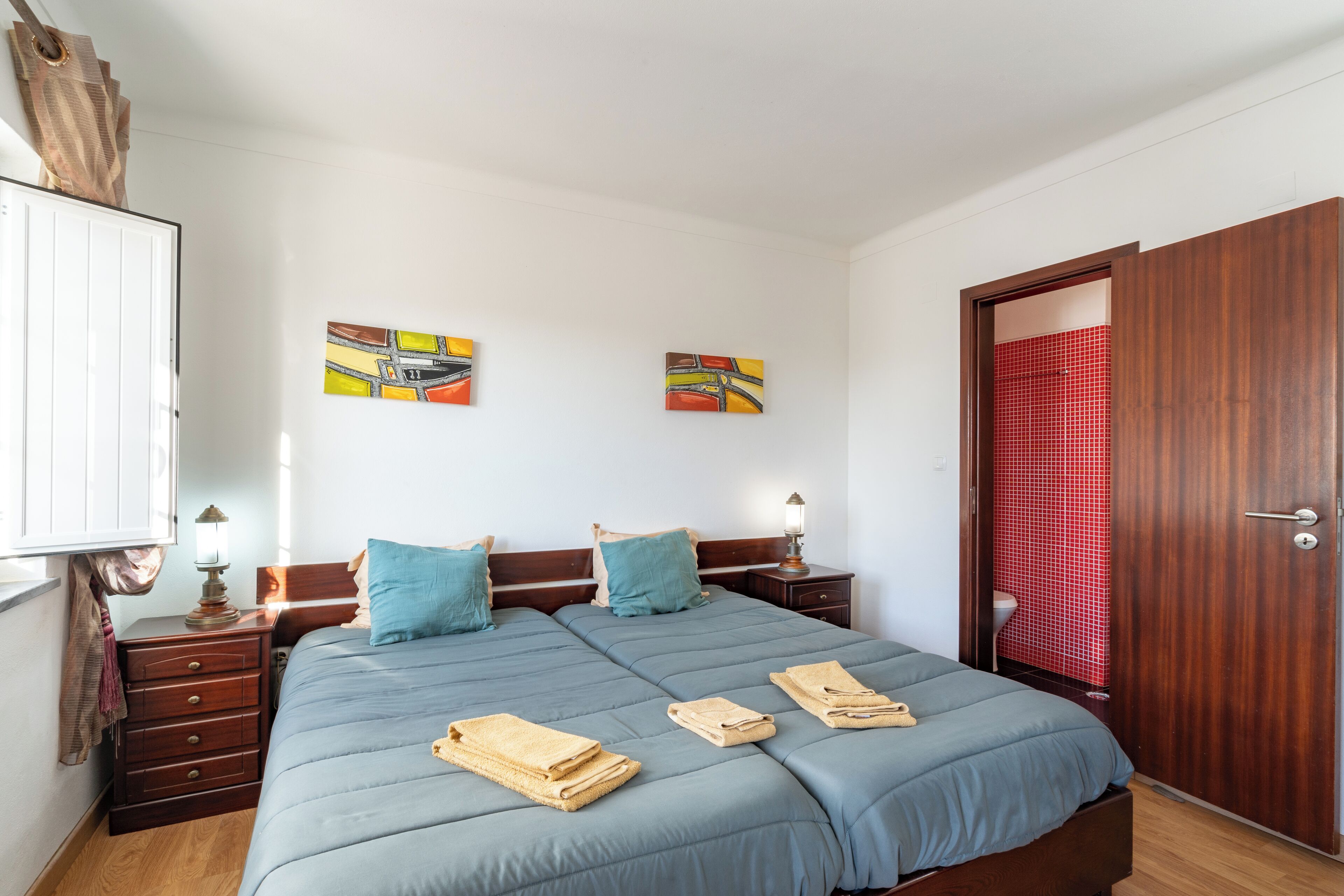 Un lit double avec une literie bleue, deux tables de chevet avec des lampes, une armoire en bois et deux œuvres d’art murales.