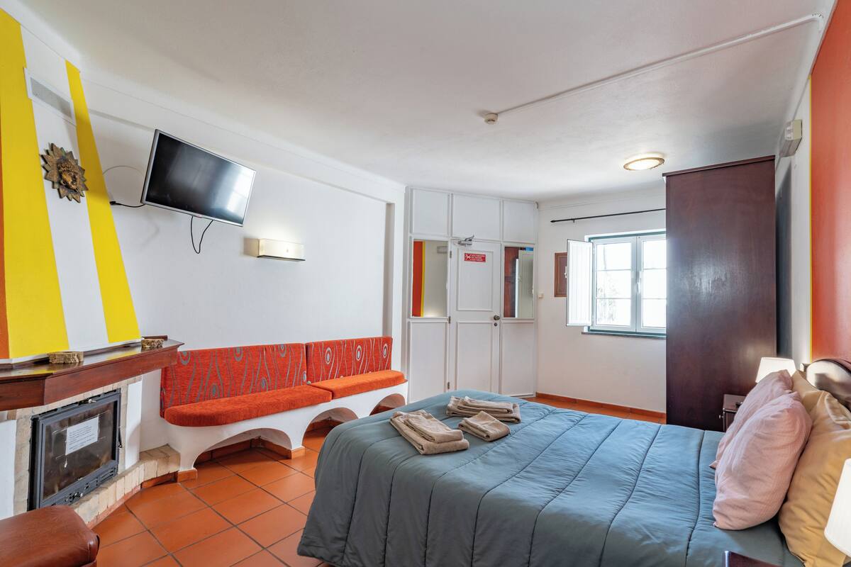 Habitación de hotel con una cama, televisión montada en la pared, chimenea, banco y puerta con espejo.