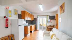 Cocina compacta con refrigerador, microondas y armarios de madera. Hay un comedor con mesa y sillas, y un sofá blanco en la sala.