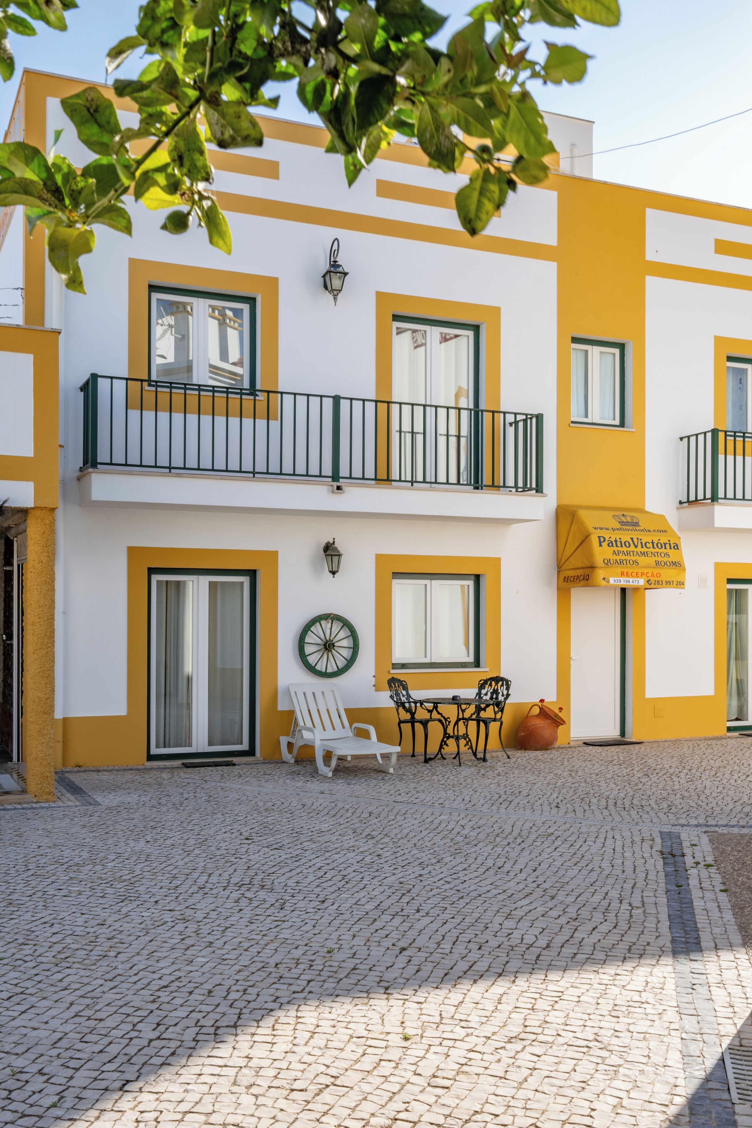 A building with yellow and white exterior walls, a balcony with a black railing, and a sign for 'Patio Victoria'.