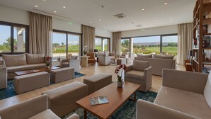 A spacious, well-lit lounge area with multiple seating options, large windows offering a view of the outdoors, and a bookshelf filled with books. - Aphrodite Hills Rentals – Junior Villas (Kouklia)