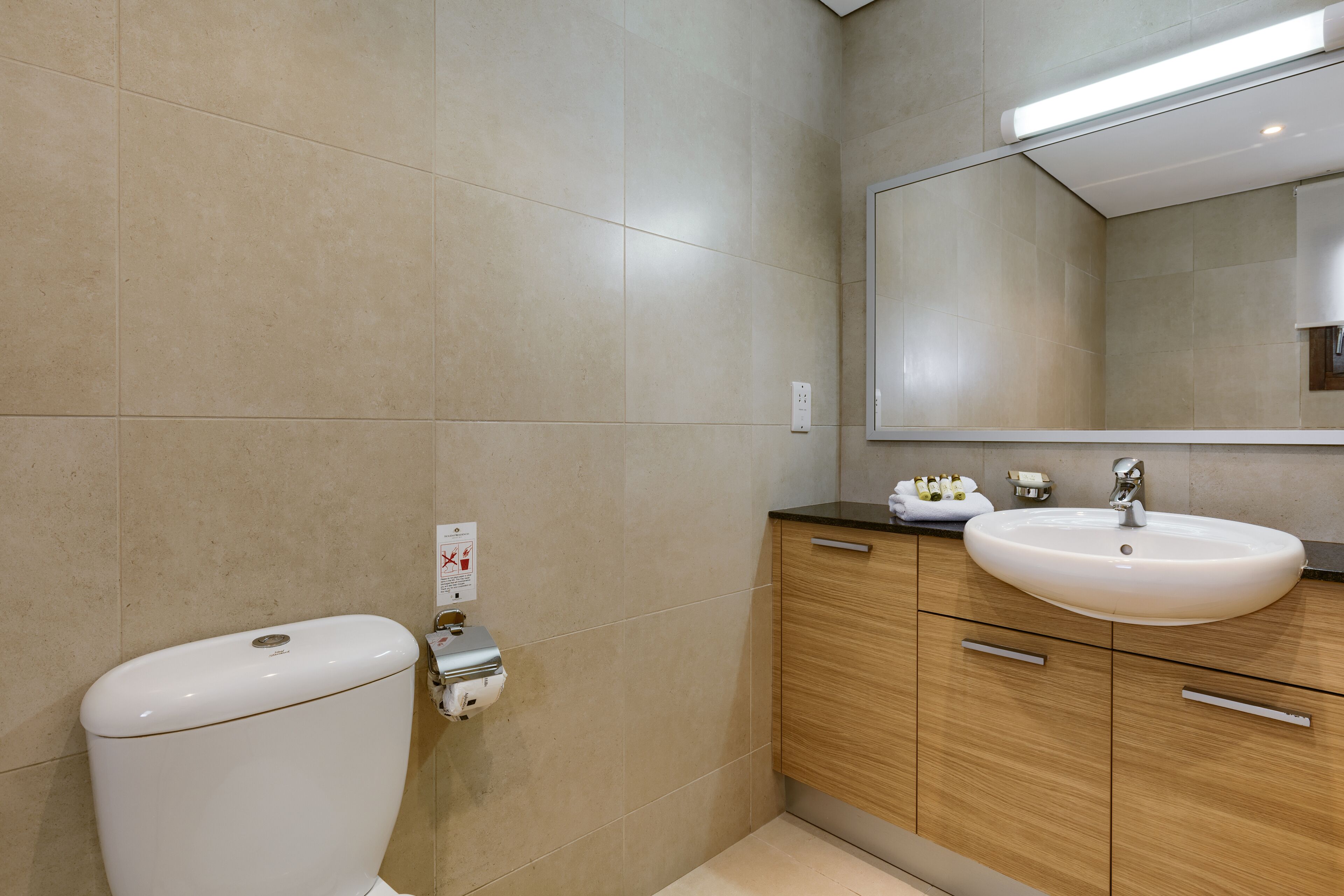 A modern bathroom with beige tiles, a white toilet, a wooden vanity with a sink, and a mirror.