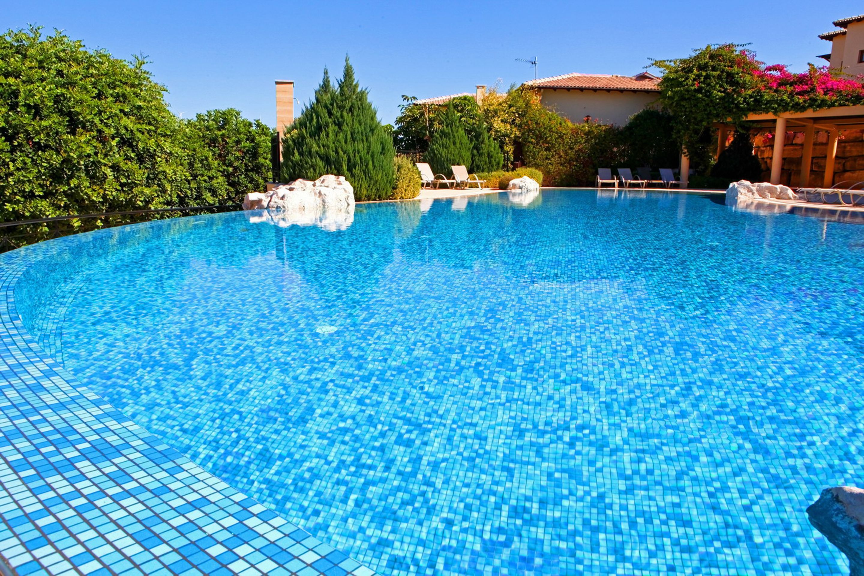 A clear blue swimming pool with tiled edges, surrounded by lush greenery and a building with a tiled roof.