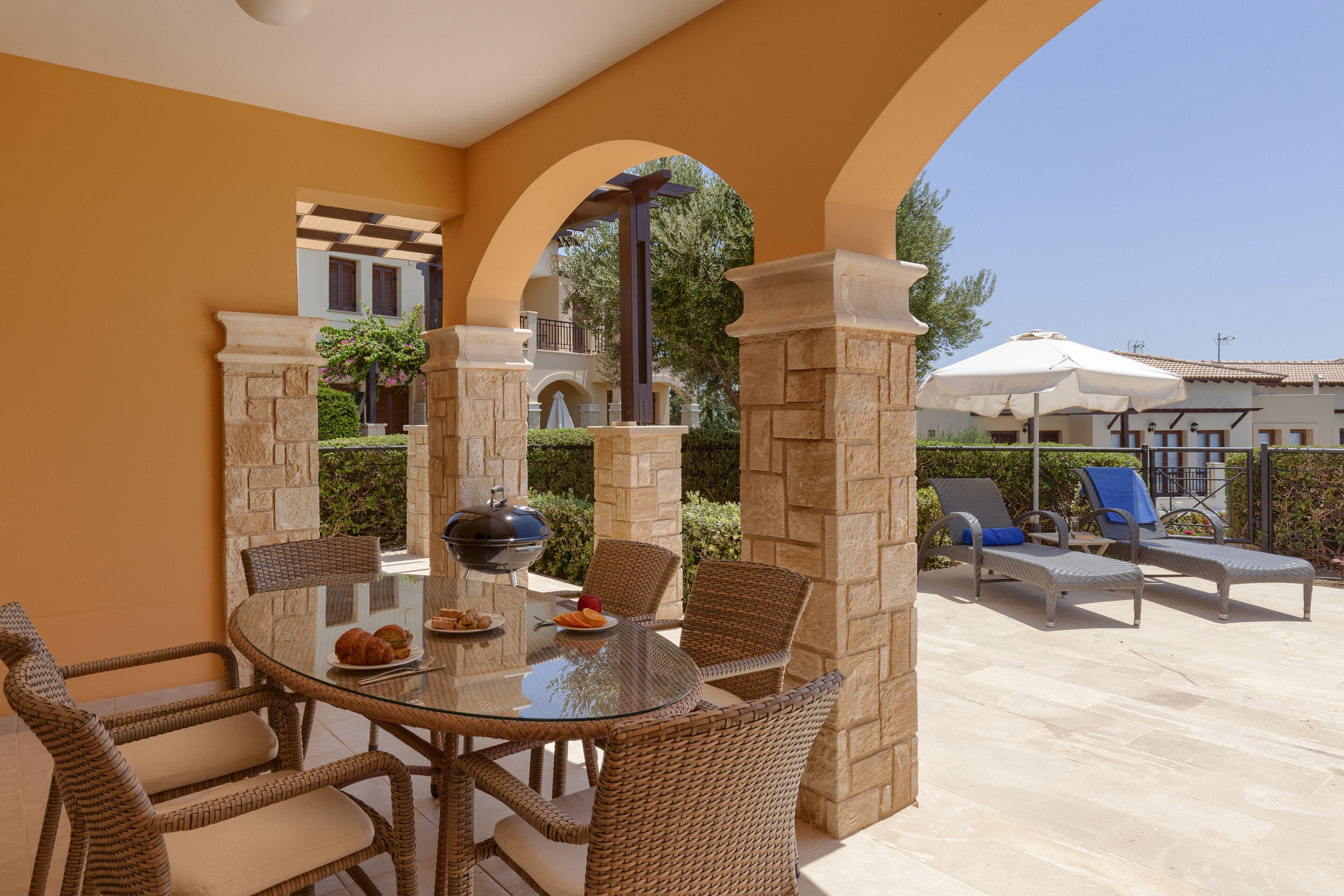 Eine Terrasse im Freien mit einem runden Glastisch, Korbstühlen und einem Grill.