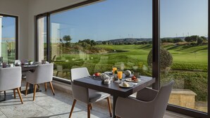 A dining area with a table set for breakfast, large windows with a view of a golf course and wind turbines. - Aphrodite Hills Rentals – Junior Villas (Kouklia)