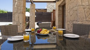 An outdoor dining area with wicker chairs, a glass table, and a stone wall backdrop. - Aphrodite Hills Rentals – Junior Villas (Kouklia)