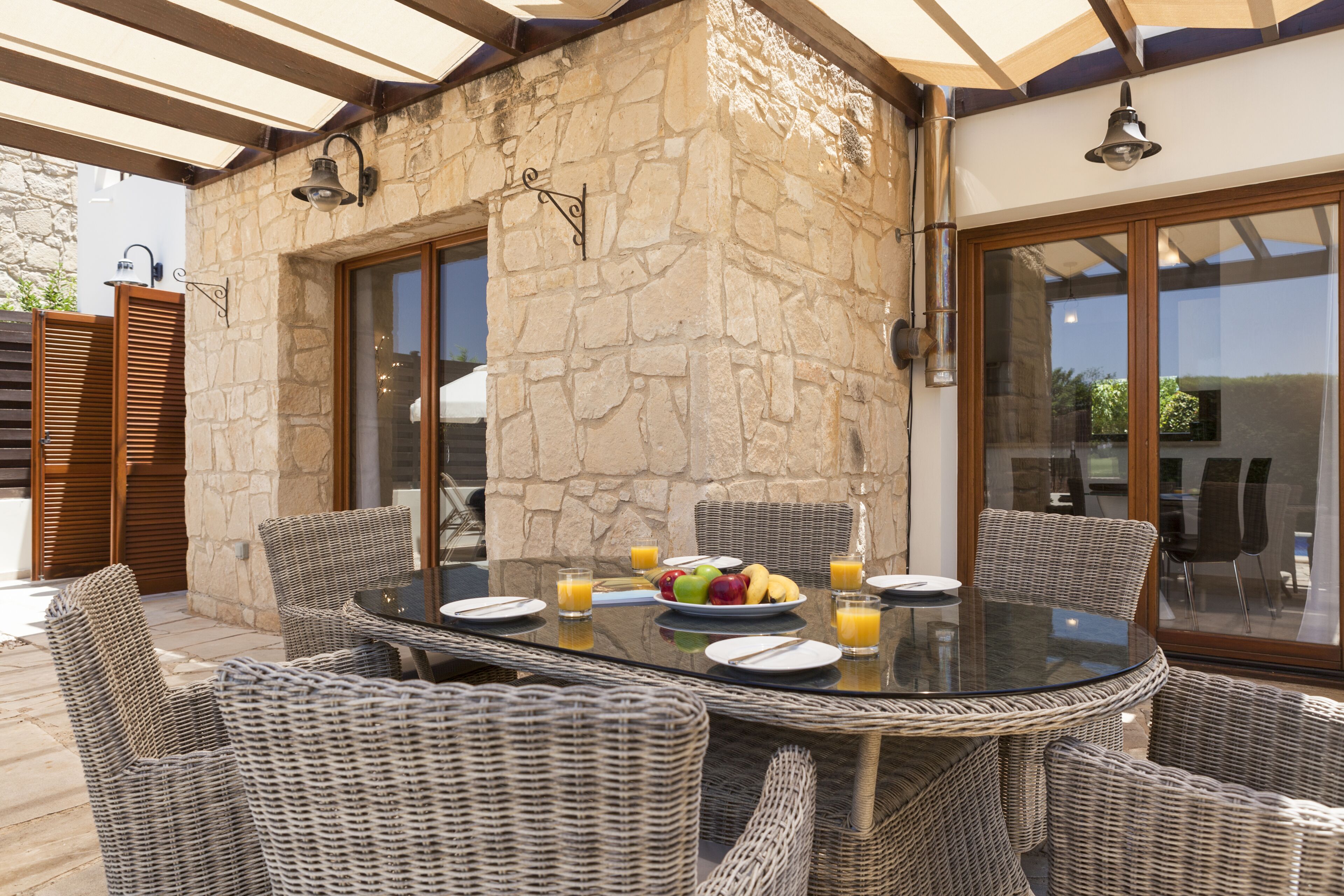 An outdoor dining area with wicker chairs and a glass table, surrounded by stone walls and large windows.
