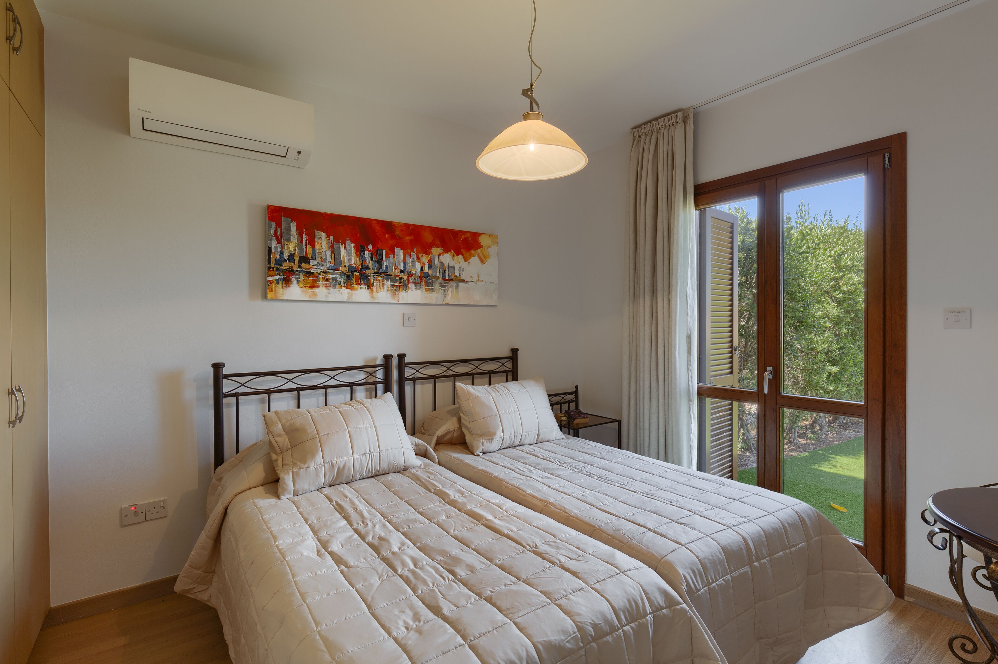 A bedroom with a bed, a painting, an air conditioner, and a window with a view of greenery.