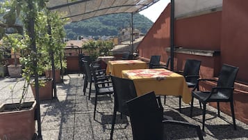 Patio esterno con tavoli e sedie, circondato da piante in vaso e affaccio su edifici sullo sfondo.