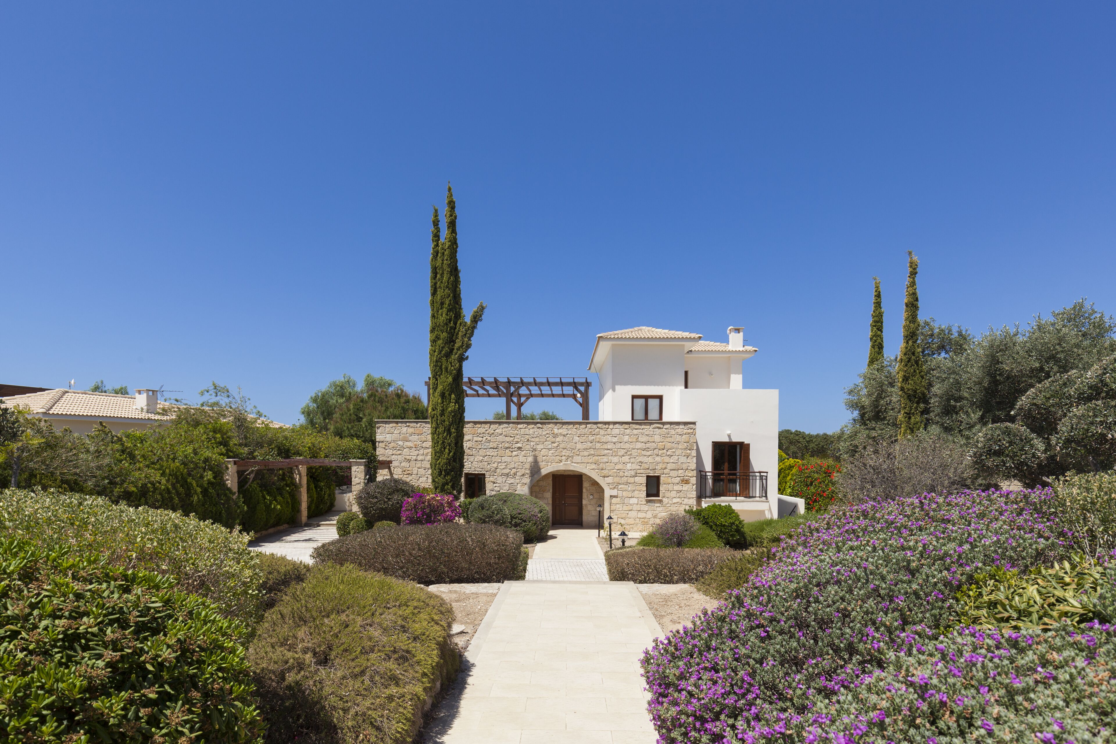 A modern house with a stone facade, a central archway, and a garden with various shrubs and flowers.
