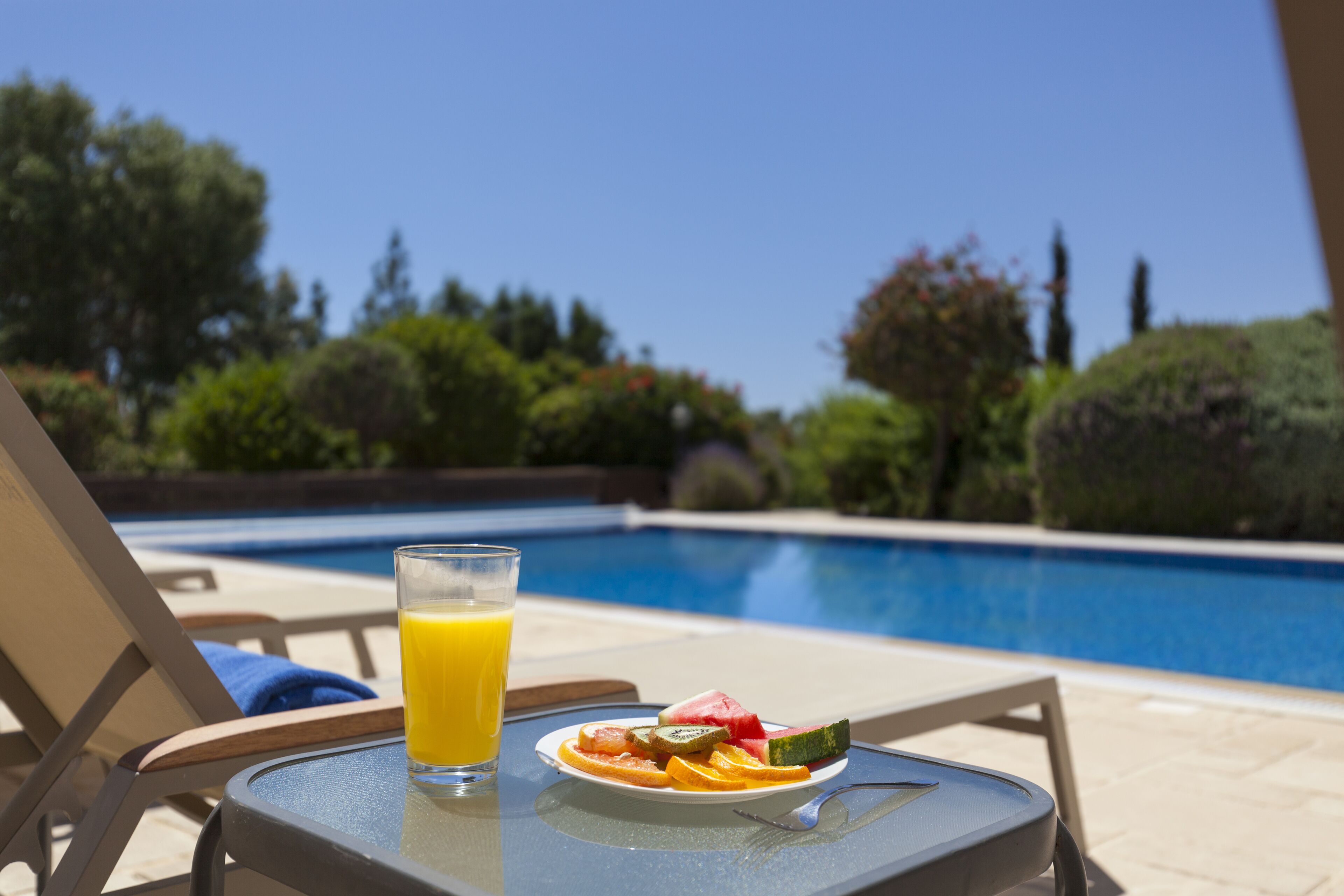 A table with a glass of orange juice and a plate of fruit near a pool.