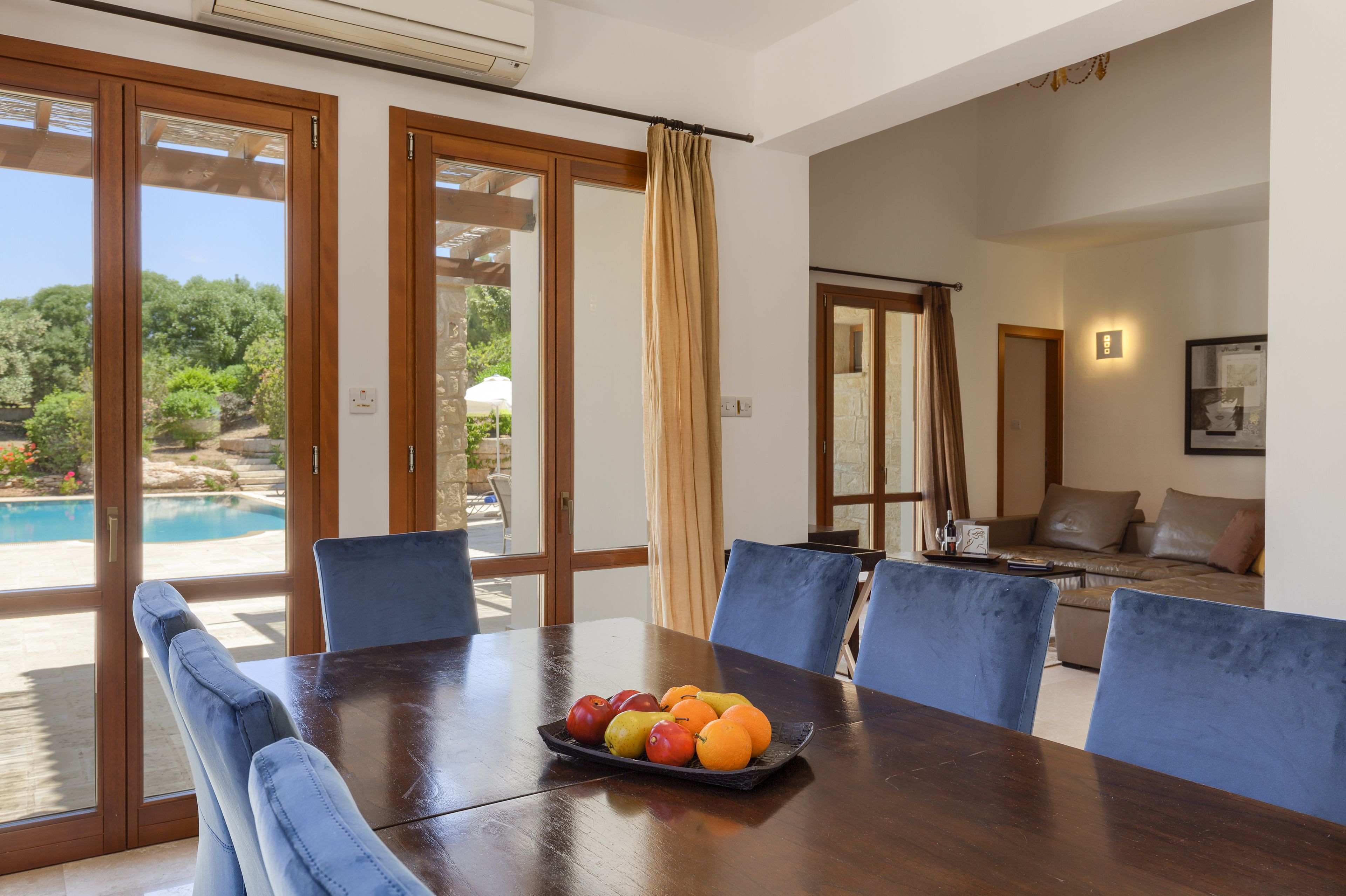 A dining area with a table set for six, a fruit bowl on the table, and a view of a swimming pool and greenery outside.