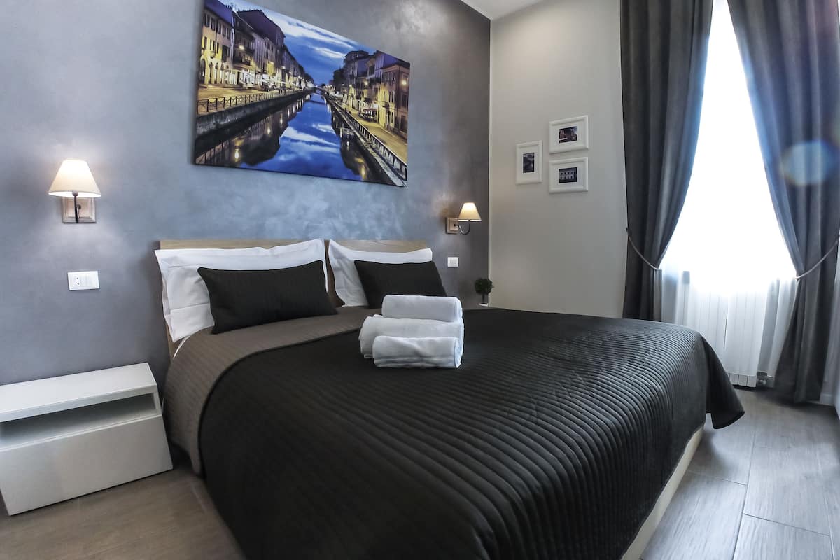 A modern hotel room with a large bed, two bedside lamps, a painting of a canal, and a window with curtains.