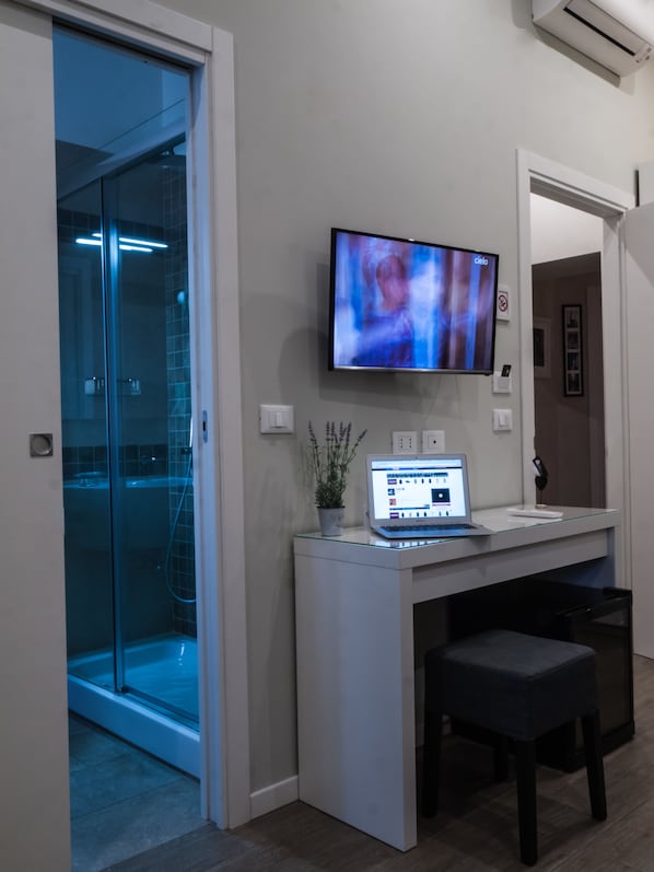 Un baño moderno con cabina de ducha de vidrio, un televisor montado y un ordenador portátil sobre un tocador de cristal.
