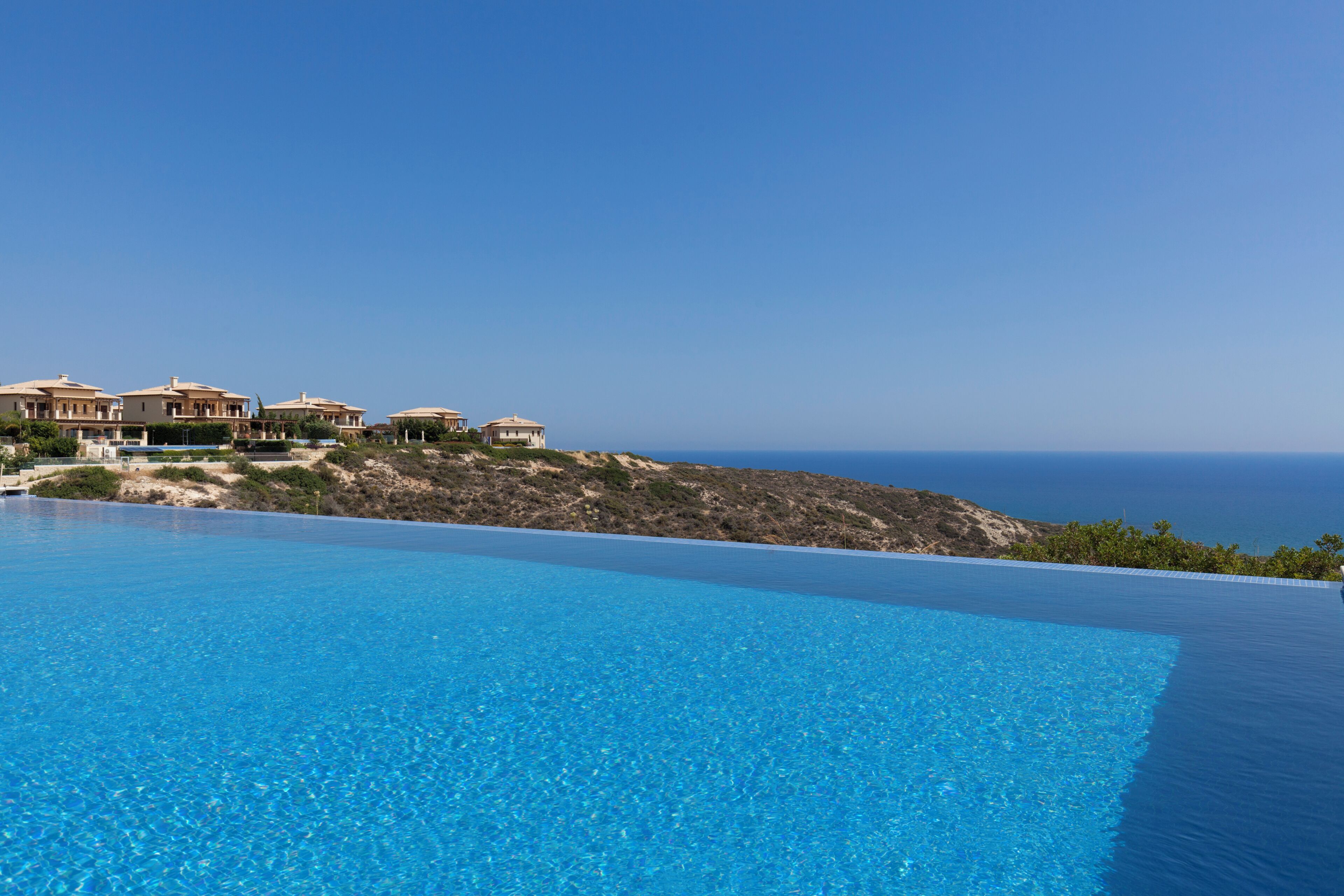 Una piscina infinita azul cristalina con vistas al océano y una colina con edificios.