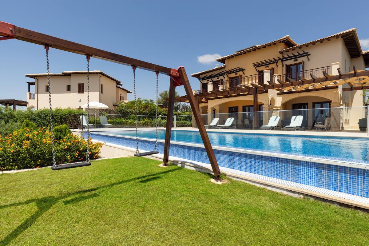 Una piscina con un columpio de madera, una zona de descanso junto a la piscina y un edificio con tejado de baldosas.