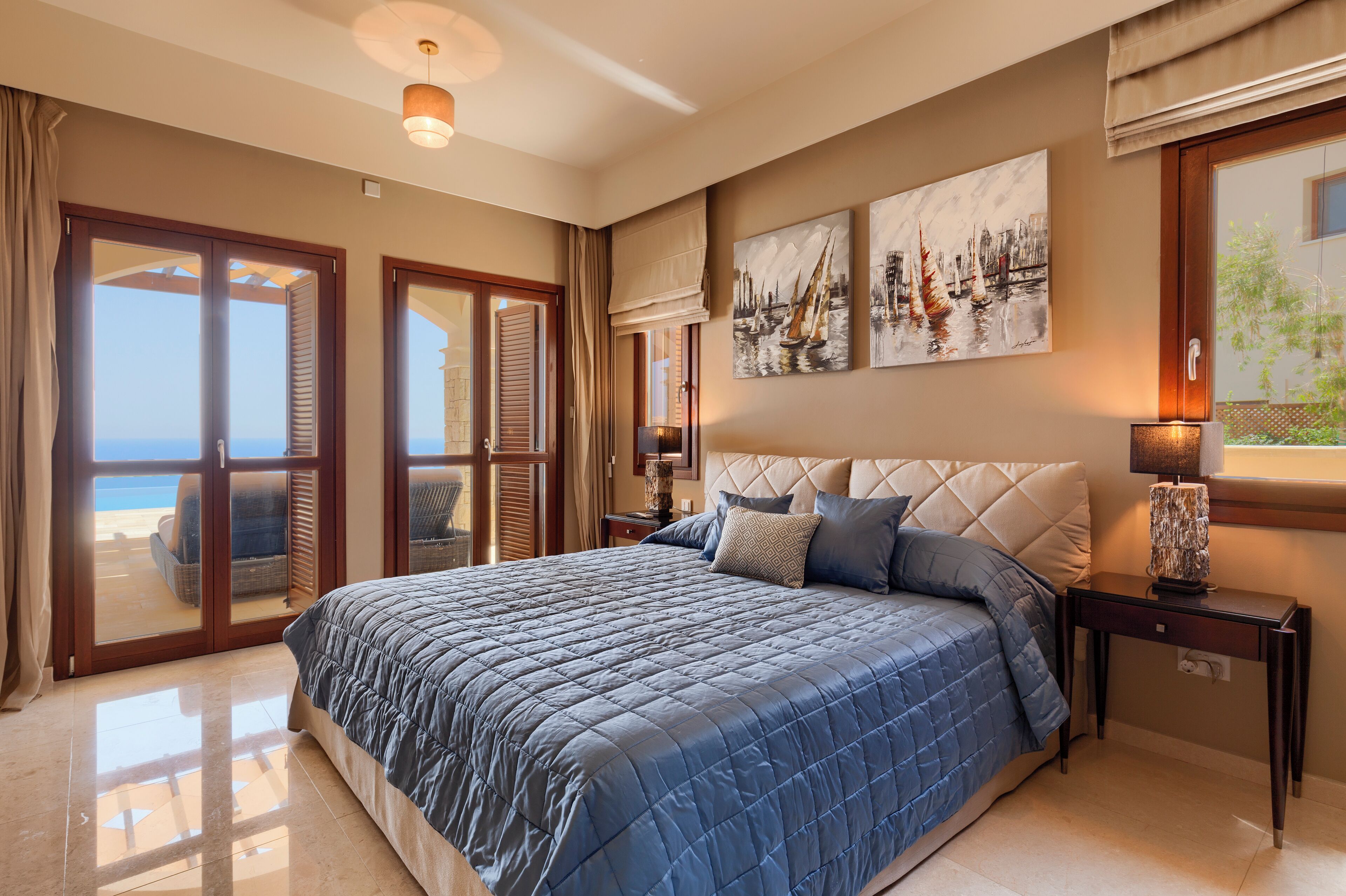 A bedroom with a large bed, a view of the sea through a sliding door, and two paintings on the wall.