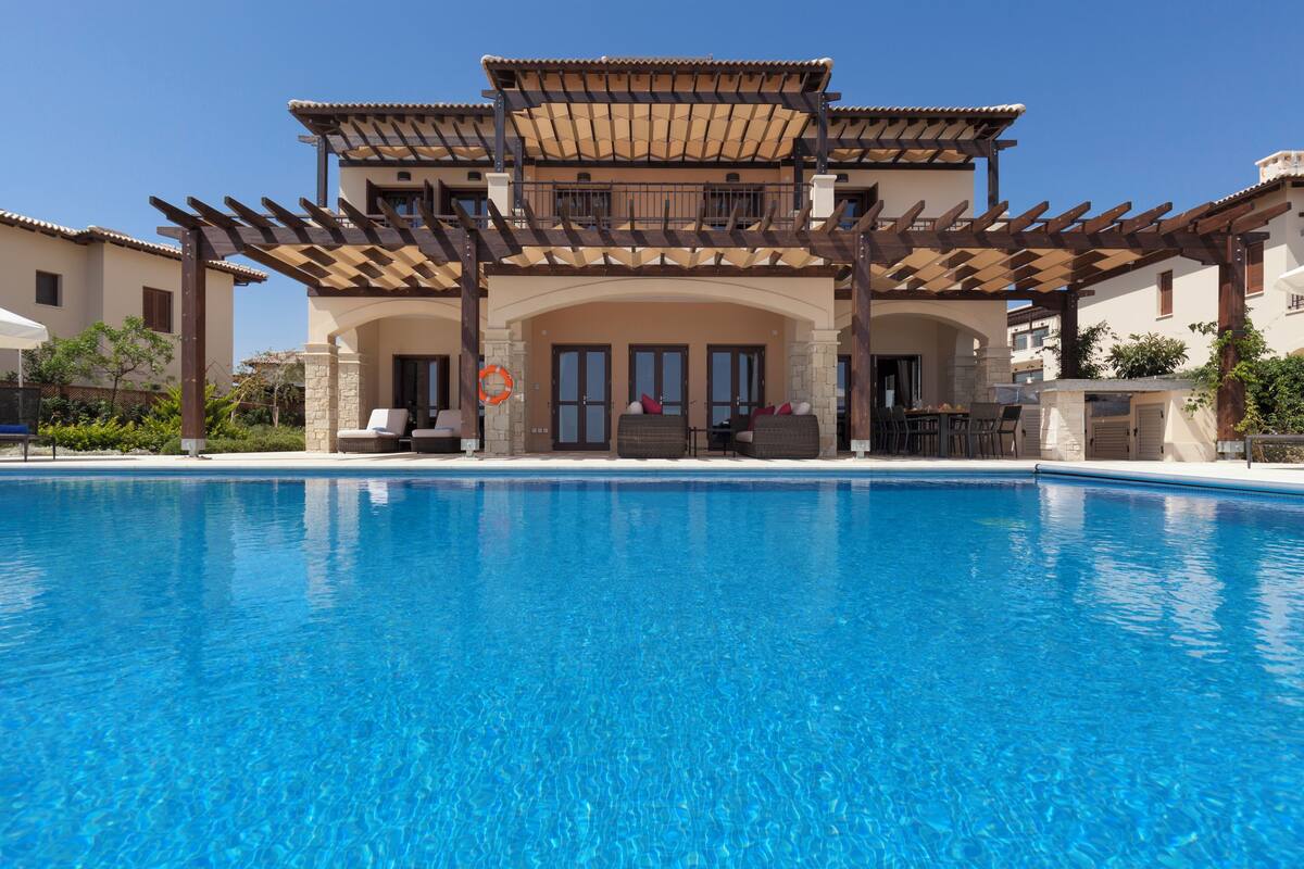 Zona junto a la piscina con una pérgola, mobiliario exterior y vistas a un edificio con tejado de baldosas.