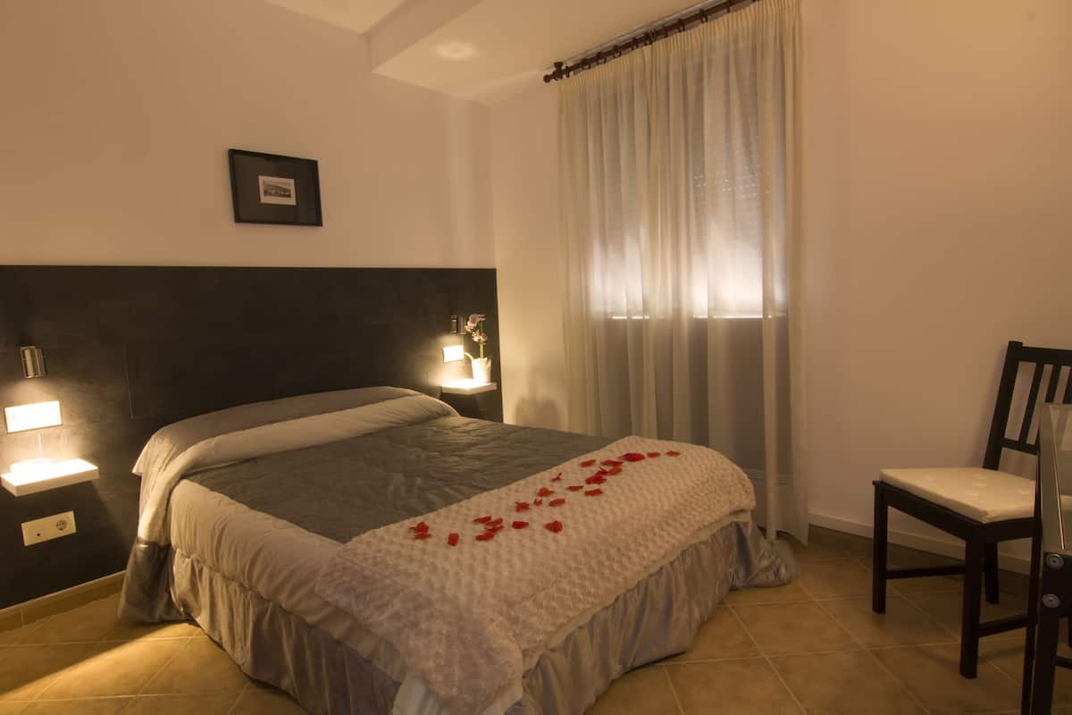Cama bem arrumada com colcha branca e arranjo decorativo de pétalas de rosa vermelha. Uma cadeira e um abajur fixado na parede estão à direita da cama.