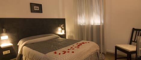 Una cama bien hecha con una colcha blanca y una decoración de pétalos de rosa roja. A la derecha de la cama hay una silla y una lámpara de pared.