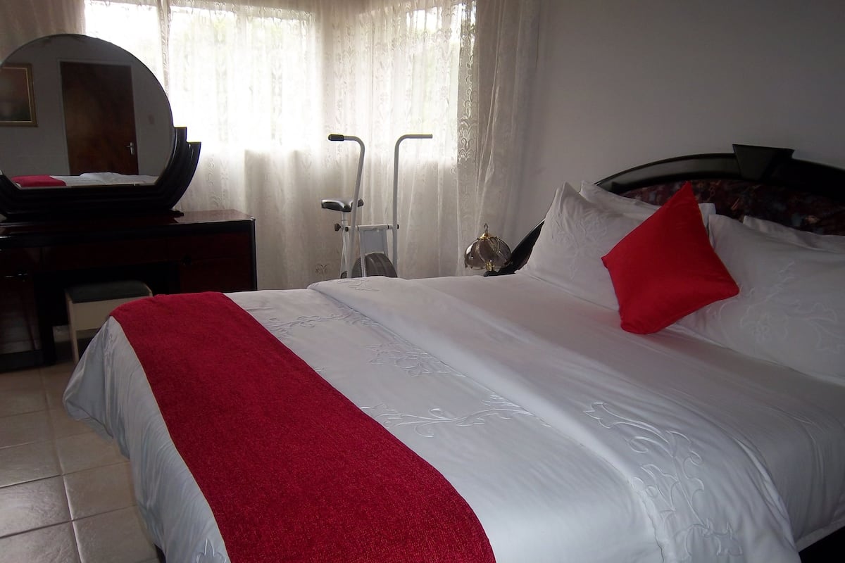 A neatly made bed with a red throw, a bedside table with a mirror, and a standing fan.