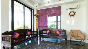 A room with two wooden sofas with cushions, a modern chair, a large window with a view, and a ceiling fan. - SakSukSmile Resort (Sukhothai)